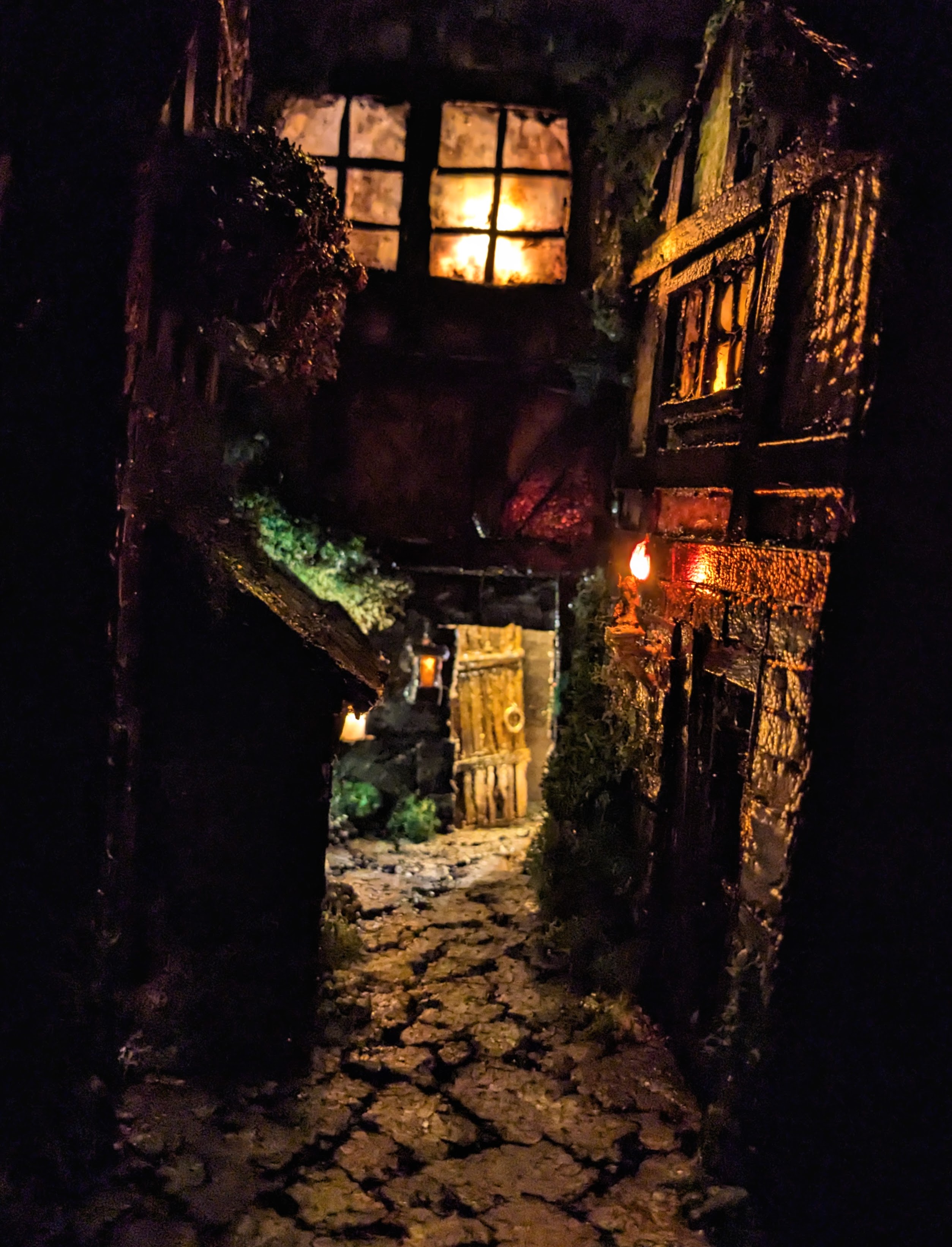 Shot of minature medieval street scene. Shows a small alley-like street with a house on either side, and another behind them against the back wall. The scene is dimly lit by a minature candle mounted on the outside of one of the houses. Light also shines out of the door of the house in the back, which is ajar.