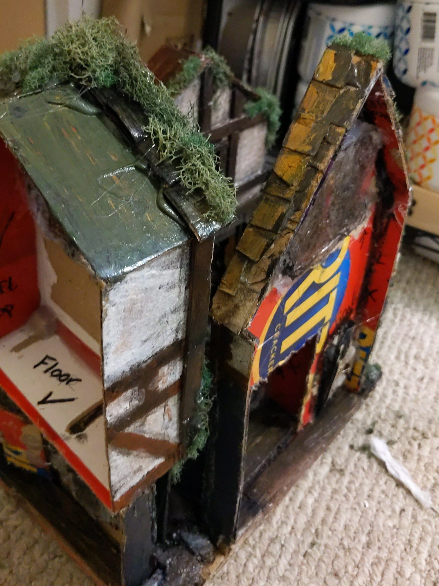 A photo showing the outside of the minature scene. You can see exposed cardboard, including a Ritz box, as well as uncovered hot glue and unfinished areas of paint. On the cardboard are traces of notes and labels.
