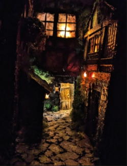 Minature medieval street scene. A small alley-like street with a house on either side, and another behind them against the back wall. Dimly lit by a minature candle on the outside of one of the houses. Light also shines out of the door of the house in the back, which is slightly open.